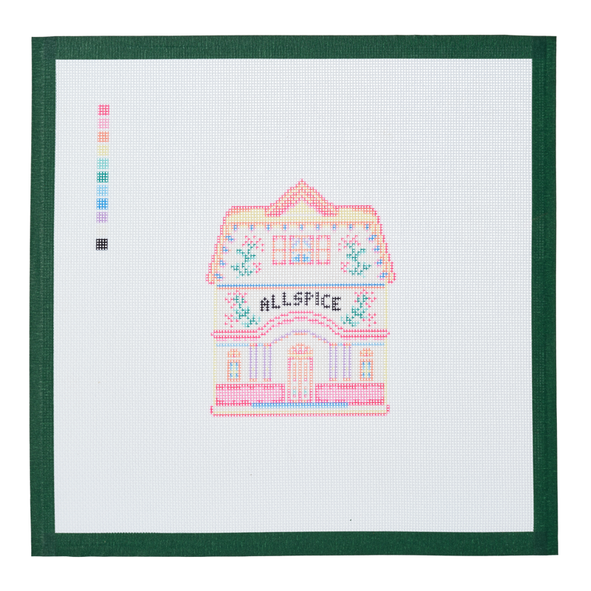 A needlepoint canvas of the Allspice house, a small pastel building with its name on the front, on white mesh with a green taped edge.