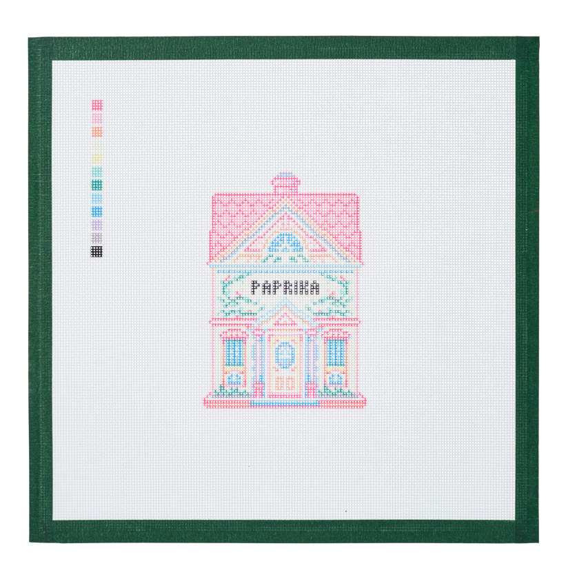 A needlepoint canvas of the Paprika house, a small pastel building with its name on the front, on white mesh with a green taped edge.