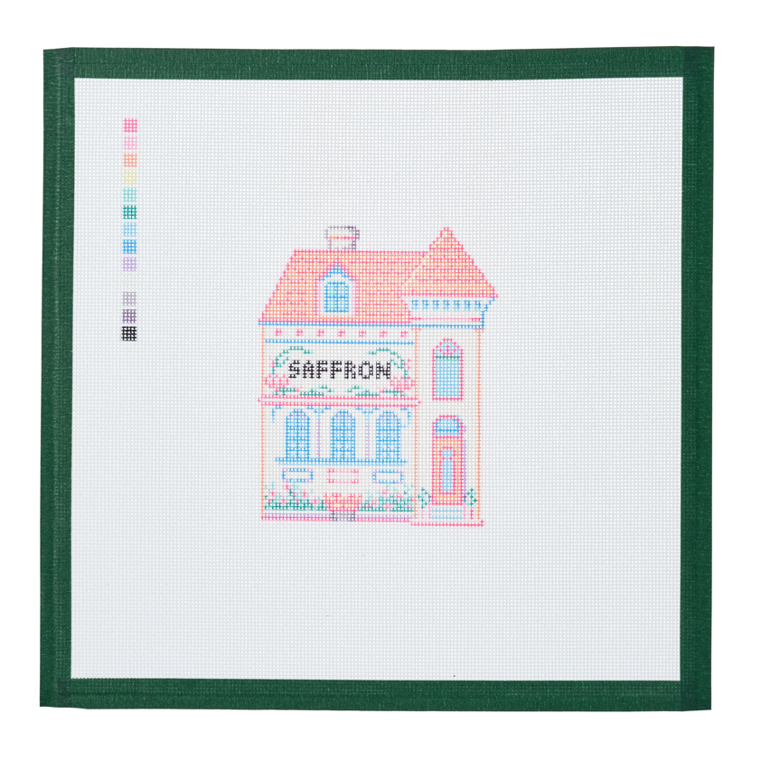 A needlepoint canvas of the Saffron house, a small pastel building with its name on the front, on white mesh with a green taped edge.