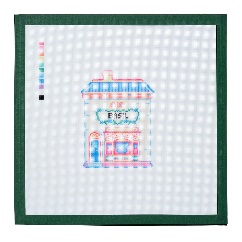A needlepoint canvas of the Basil house, a small pastel building with its name on the front, on white mesh with a green taped edge.