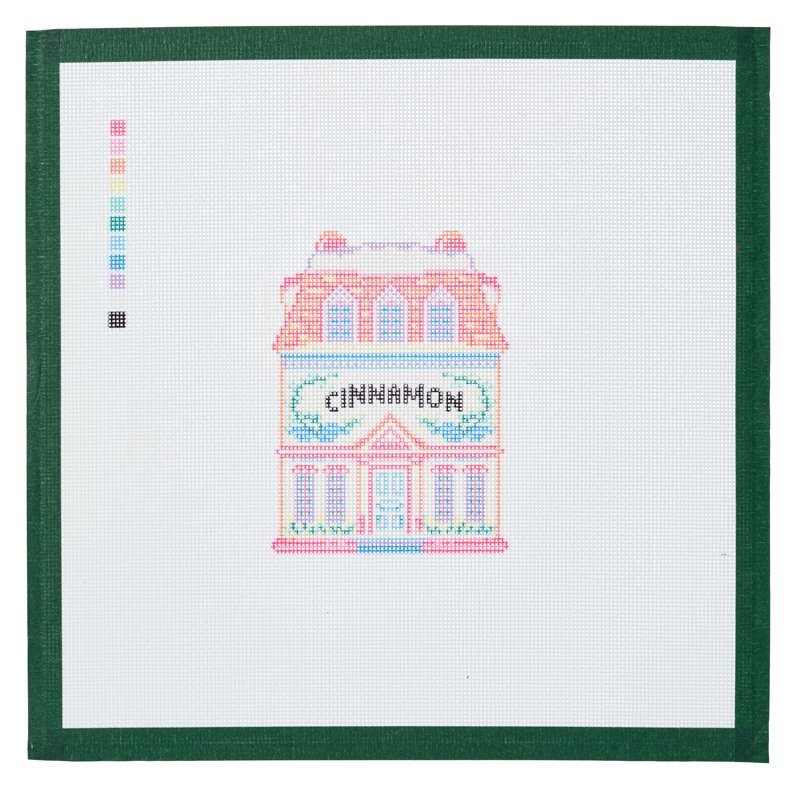 A needlepoint canvas of the Cinnamon house, a small pastel building with its name on the front, on white mesh with a green taped edge.