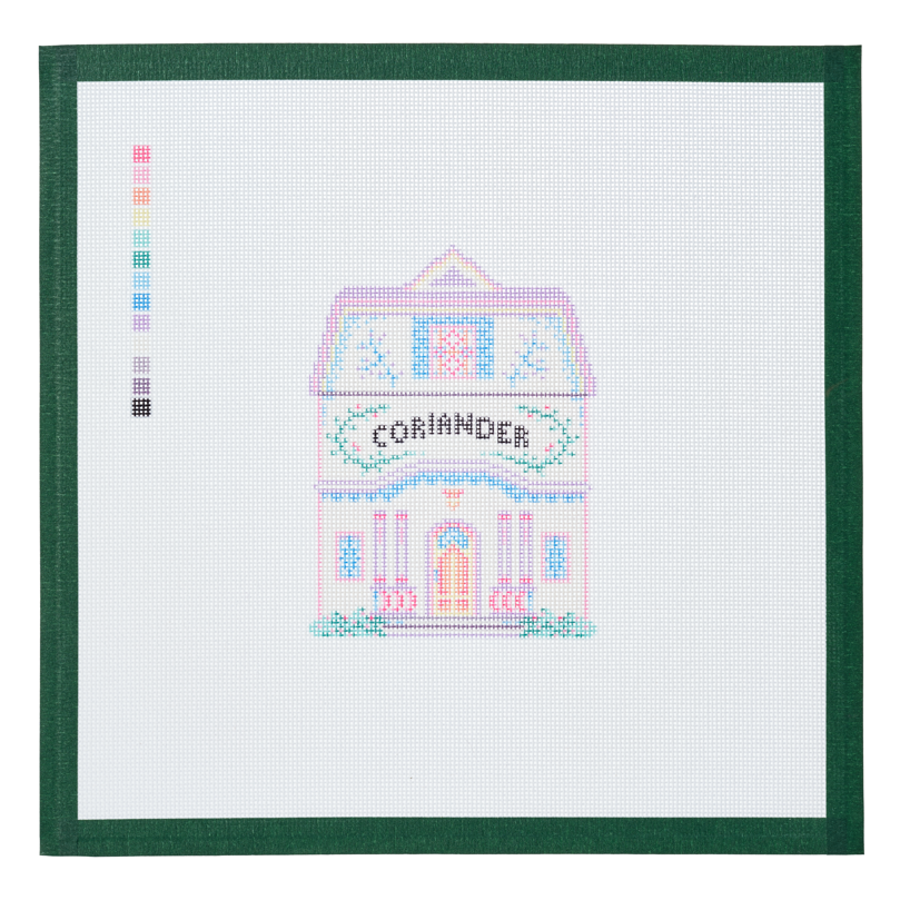 A needlepoint canvas of the Coriander house, a small pastel building with its name on the front, on white mesh with a green taped edge.