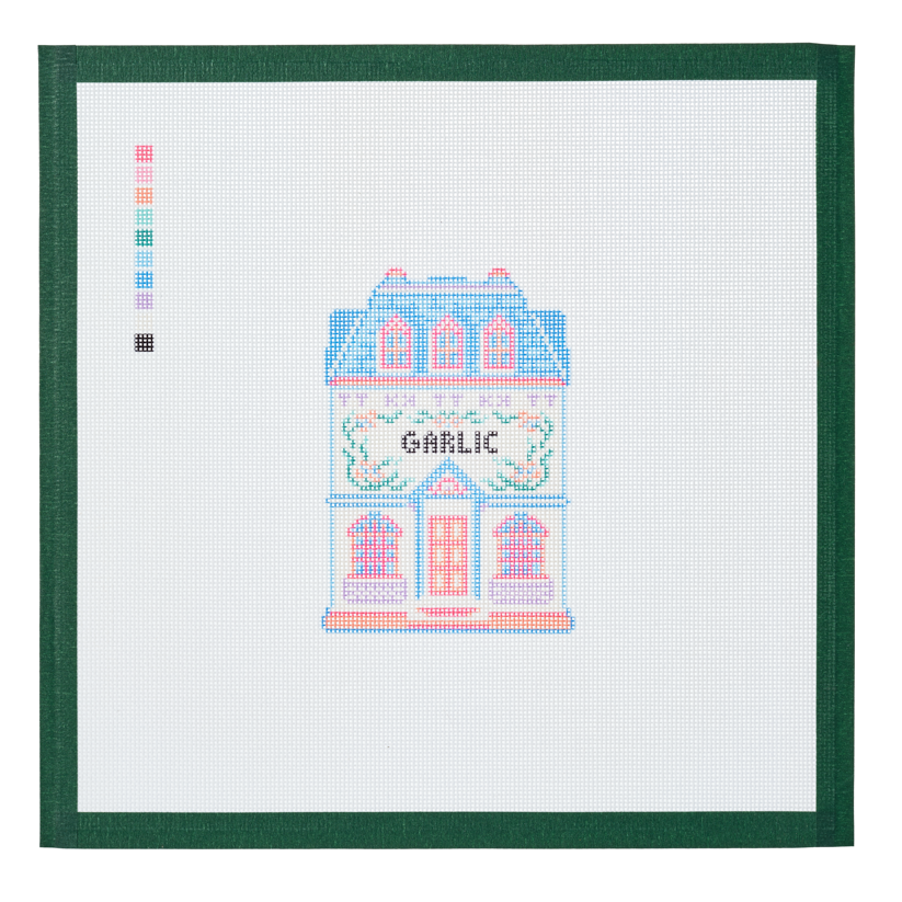 A needlepoint canvas of the Garlic house, a small pastel building with its name on the front, on white mesh with a green taped edge.