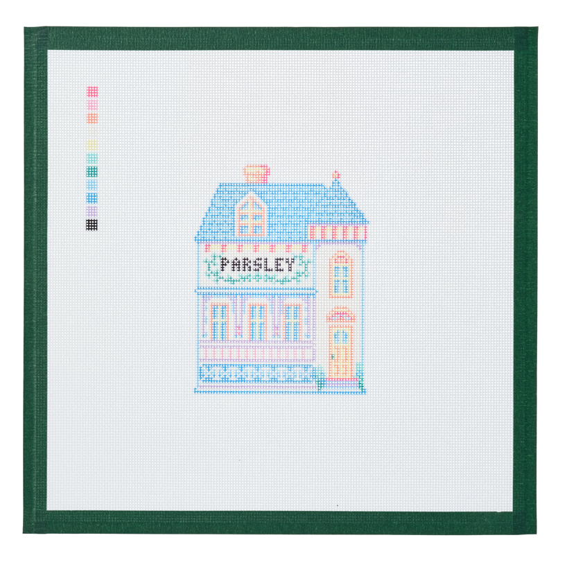 A needlepoint canvas of the Parsley house, a small pastel building with its name on the front, on white mesh with a green taped edge.