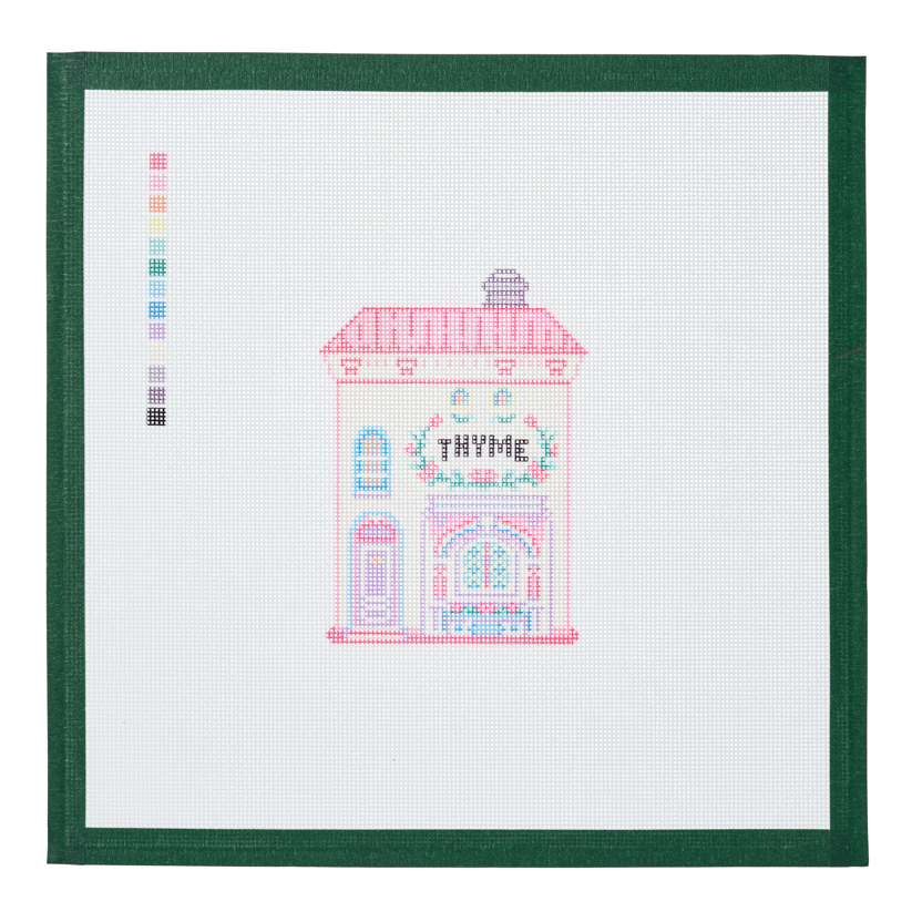 A needlepoint canvas of the Thyme house, a small pastel building with its name on the front, on white mesh with a green taped edge.